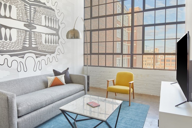 Modern loft living room with gray couch, yellow accent chair, marble and metal coffee table, large modern floor lamp, wall mural, industrial windows.